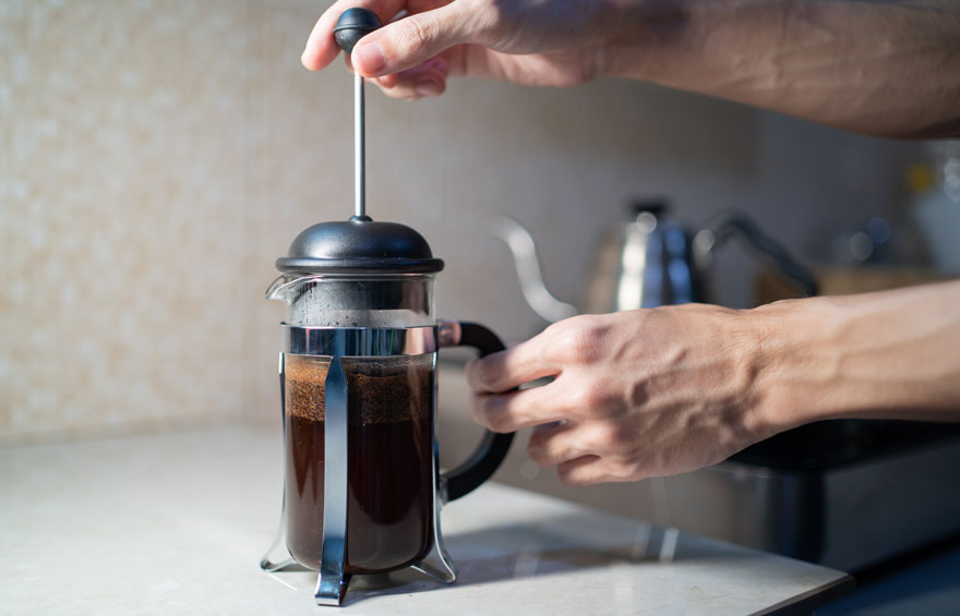 Cafetière en verre avec café, couvercle noir et piston enfoncé par une main sur un plan de travail clair. Bouilloire en arrière-plan.