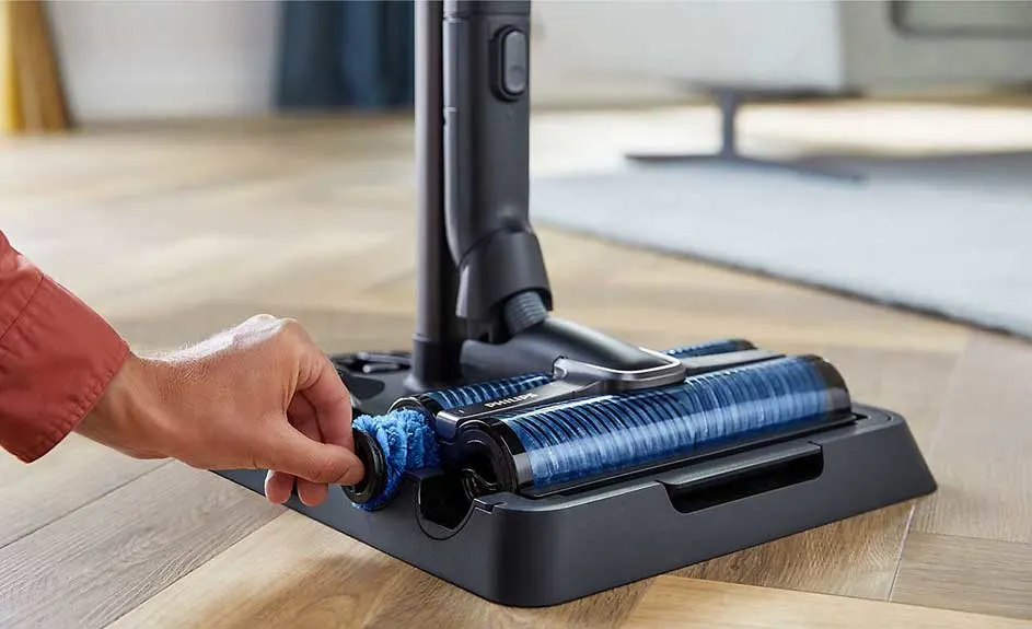 Main d'une personne ajustant une brosse bleue sur une base noire d'aspirateur sur un sol en bois clair.