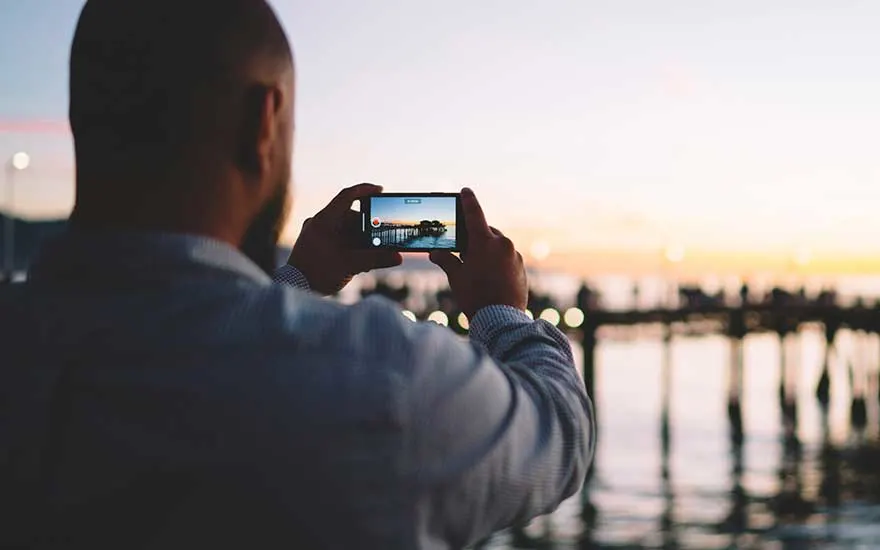 A man takes a photo of a pier at sunset with his smartphone, as evening lights reflect softly across the water’s surface.