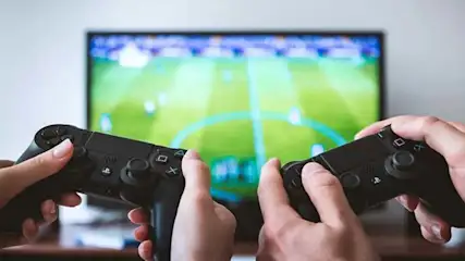 Two hands hold black game controllers in front of a screen showing a green field.