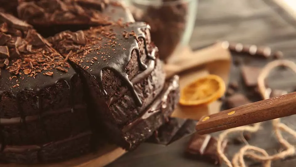 Stapel chocoladebrownies met glazuur en schaafsel, naast een schijfje sinaasappel en een houten mes op een rustieke ondergrond.