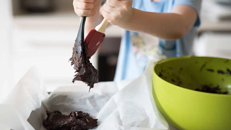 Pâtissier étalant de la pâte à brownie au chocolat avec une spatule dans un plat à pâtisserie garni de papier sulfurisé. Bol vert à côté.