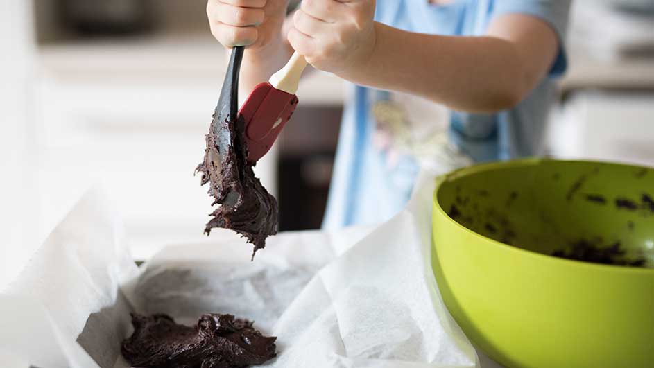 Een spatel verdeelt bruin deeg in een bakvorm met bakpapier, naast een groene kom.