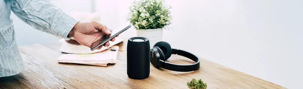 Main tenant un téléphone près d'un haut-parleur noir, d'un casque, de papiers et d'une plante sur une table en bois.