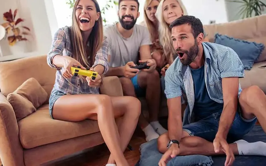 Vier vrienden zitten in een gezellige woonkamer, spelen videogames met controllers, zittend op een bank en kussens op de grond.