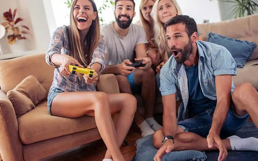 Four friends are sitting in a cosy living room, playing video games with controllers while seated on a sofa and cushions on the floor.