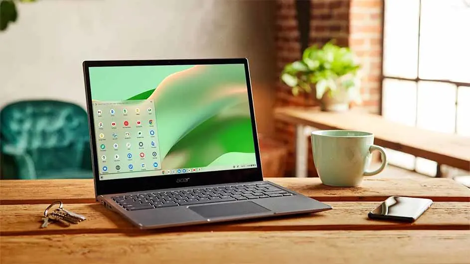 Een laptop staat op een houten tafel met een kop, telefoon en sleutels. Op de achtergrond een bakstenen muur en plant.