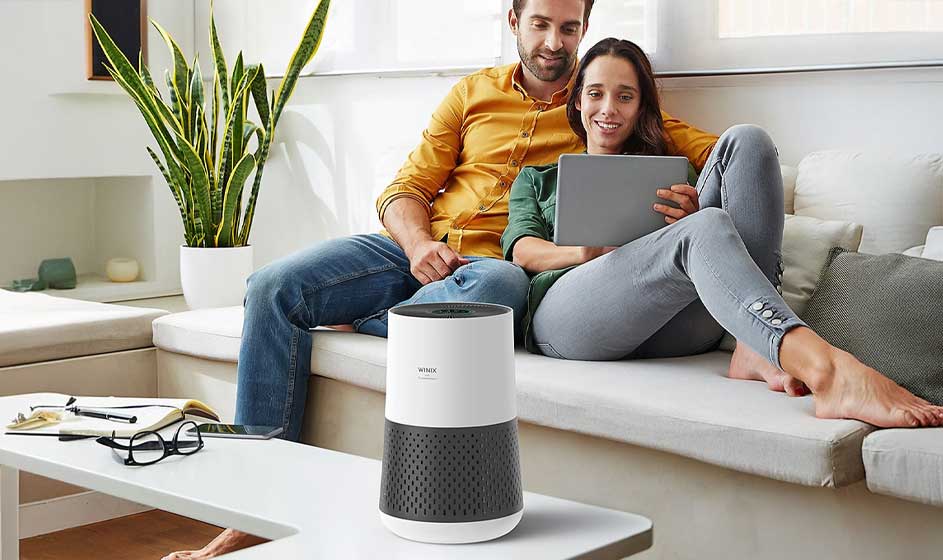 Un couple se détend sur un canapé clair en regardant une tablette, tandis qu’au premier plan un petit purificateur d’air blanc assainit la pièce.