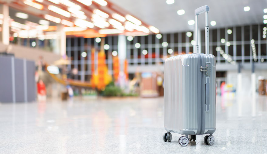 Valise argentée à roulettes dans un hall spacieux avec des drapeaux orange et des lumières vives au plafond.