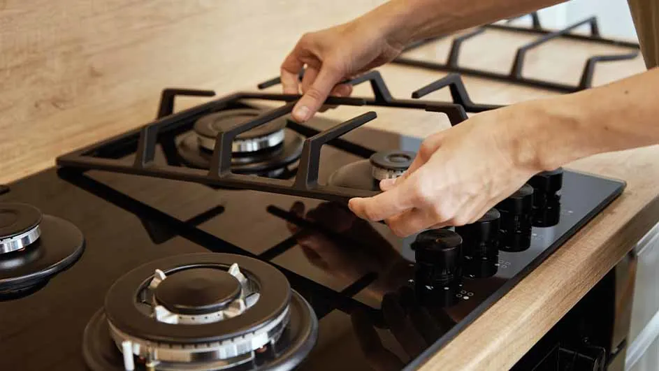 Une personne place une grille noire sur une cuisinière à gaz noire avec des boutons de commande noirs et un comptoir en bois clair.