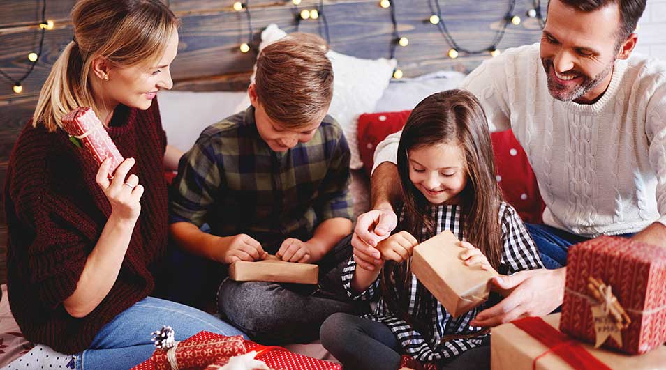 Une famille est confortablement réunie sur le canapé, ouvrant joyeusement des cadeaux de Noël, entourée de guirlandes lumineuses.