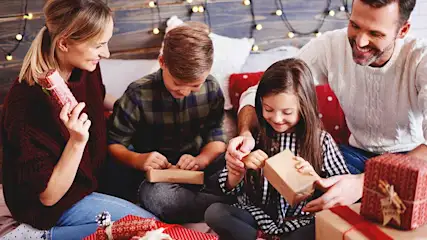 Une famille est confortablement réunie sur le canapé, ouvrant joyeusement des cadeaux de Noël, entourée de guirlandes lumineuses.