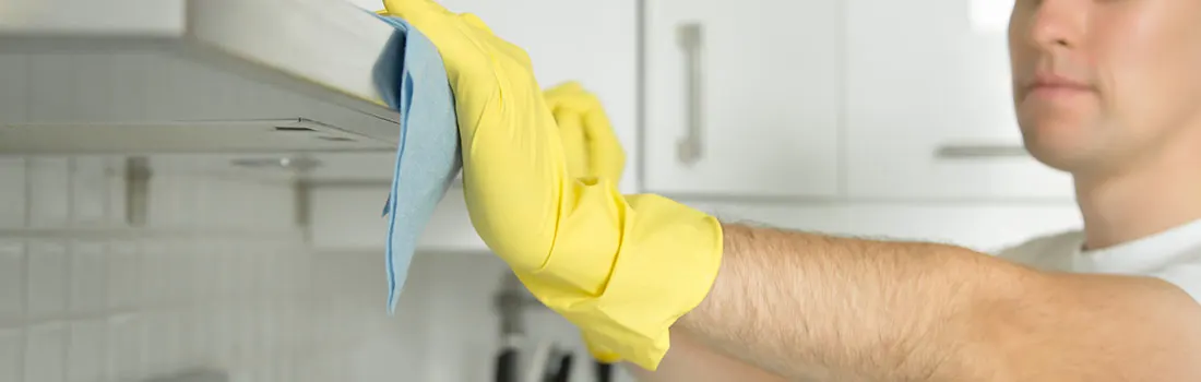 Une main gantée de jaune nettoie une hotte grise avec un chiffon bleu dans une cuisine blanche.