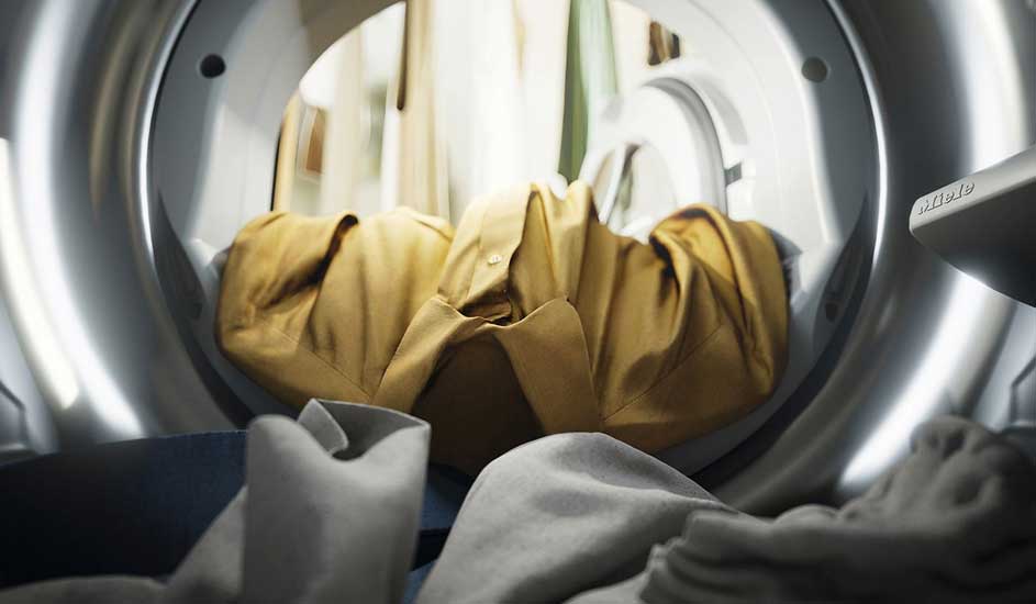 L'intérieur d'un sèche-linge avec un tambour métallique, contenant des vêtements, dont une chemise jaune et d'autres tissus dans des tons doux.