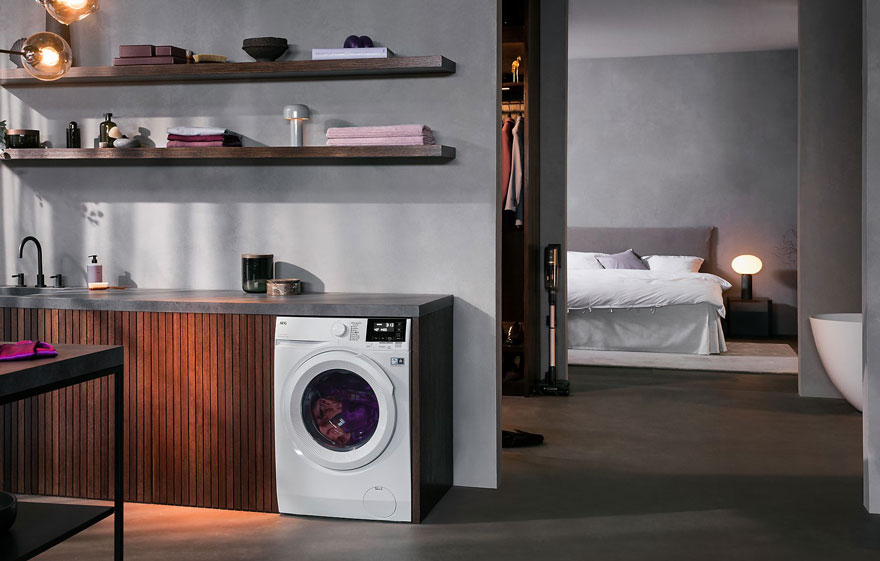 Witte wasmachine naast een donkerhouten kast en een aanrecht onder houten planken aan een grijze muur. Slaapkamer op achtergrond.