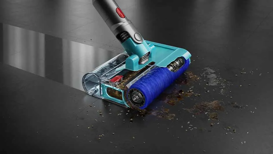 Aspirateur turquoise et argenté sur un sol sombre avec des débris. La tête de l'aspirateur est transparente.