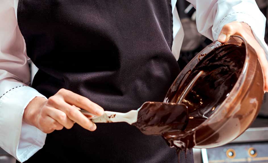 Préparation de chocolat : un bol marron rempli de chocolat fondu est incliné, une spatule blanche mélange. Tablier noir visible.