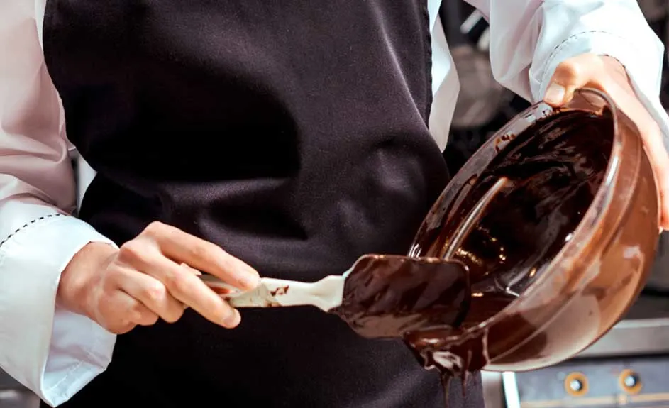 Préparation de chocolat : un bol marron rempli de chocolat fondu est incliné, une spatule blanche mélange. Tablier noir visible.