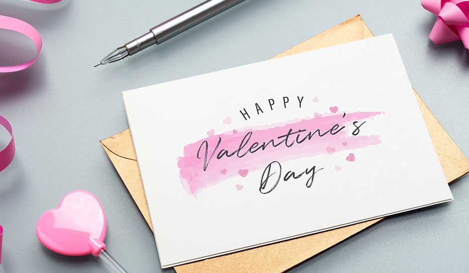 Carte de Saint-Valentin blanche sur enveloppe beige, avec stylo argenté et décorations roses sur fond gris.