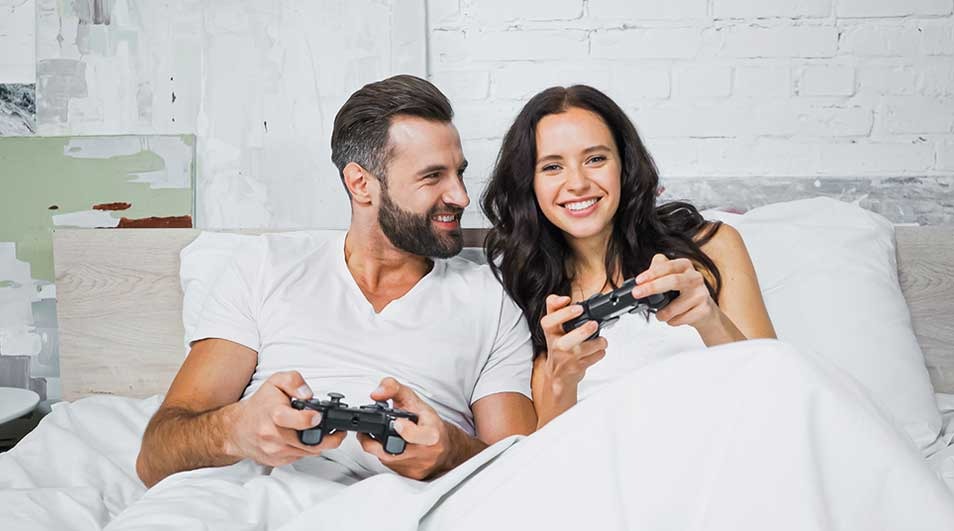 Een lachend koppel ligt samen in bed in witte lakens, ieder met een gamecontroller, ontspannen spelend in een lichte slaapkamer