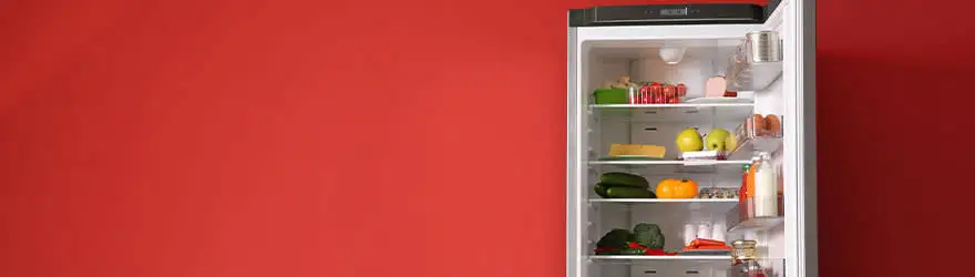 Un réfrigérateur ouvert, rempli de fruits, de légumes et de boissons, se trouve devant un mur rouge uni.