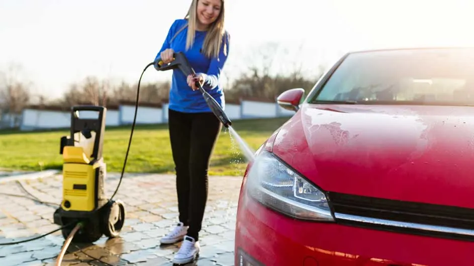 Une voiture rouge est nettoyée avec un nettoyeur haute pression jaune sur une allée pavée. Gazon et arbres en arrière-plan.