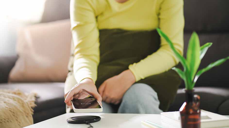 Sur une table blanche, une personne place un téléphone sur un chargeur sans fil. En arrière-plan, un canapé, des coussins et une plante verte.