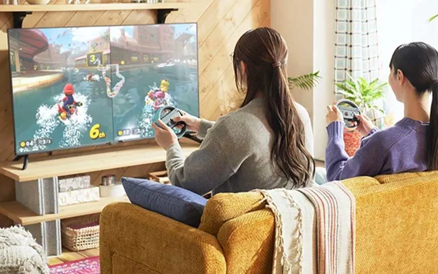 Twee personen spelen een videospel op een grote tv in een woonkamer met houten vloer en mosterdgele zetel.