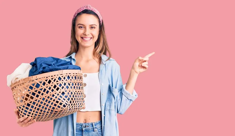 Une femme souriante tient un panier à linge en osier et pointe vers la droite sur un fond rose.