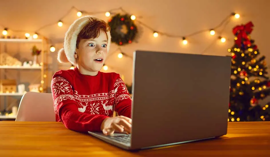 Een laptop staat op een houten tafel, met op de achtergrond kerstversiering zoals lichtjes en een kerstboom.