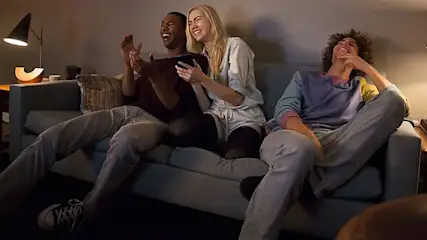 Drie mensen zitten op een grijze bank in een kamer. Een lamp staat op een kastje.