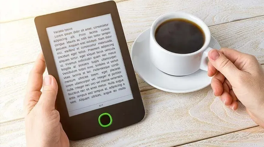 An e-reader with text is held next to a white cup of coffee on a saucer, set on a light wooden table.