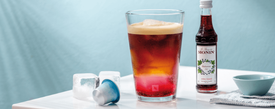 Un café glacé en couches avec des glaçons dans un verre, à côté d’une bouteille de sirop Monin, de deux glaçons et d’une capsule de café bleue sur une table blanche.
