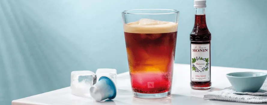 Un café glacé en couches avec des glaçons dans un verre, à côté d’une bouteille de sirop Monin, de deux glaçons et d’une capsule de café bleue sur une table blanche.