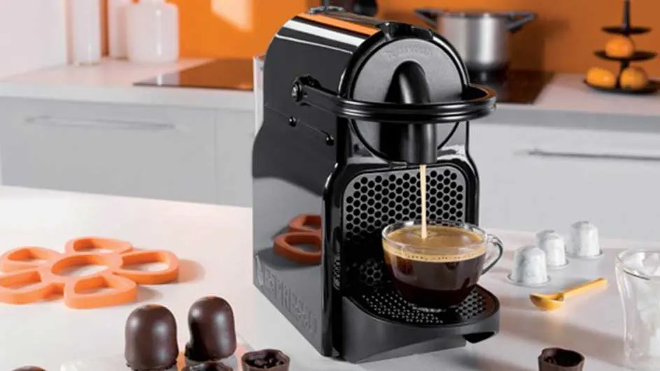 Black coffee machine pouring coffee into a glass cup. Orange coasters and sweets on a white countertop.
