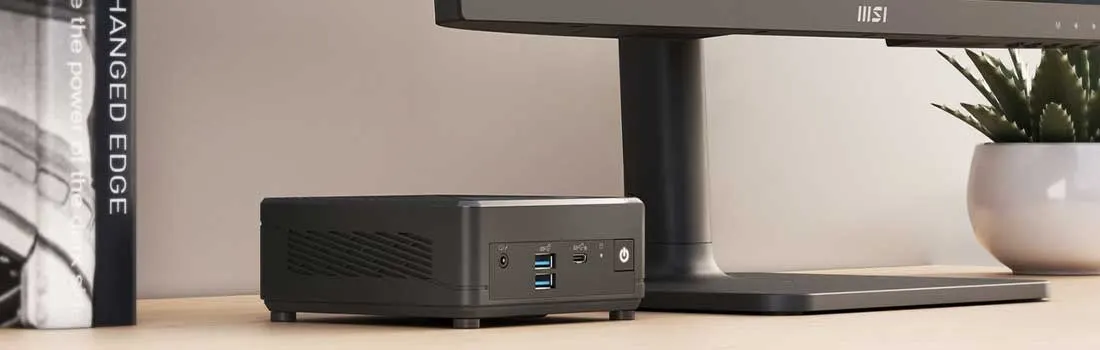 Un mini-ordinateur noir est placé sur une table en bois, avec un écran, une plante en pot et un livre en arrière-plan.