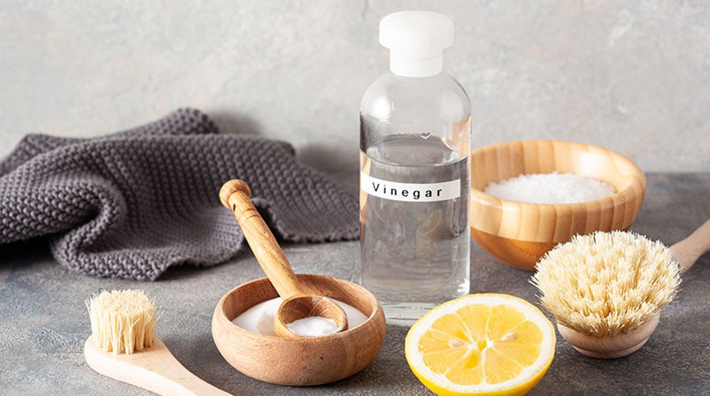 Une bouteille de vinaigre à côté d'un citron, de brosses, d'un linge gris et de bols en bois remplis de poudre blanche.