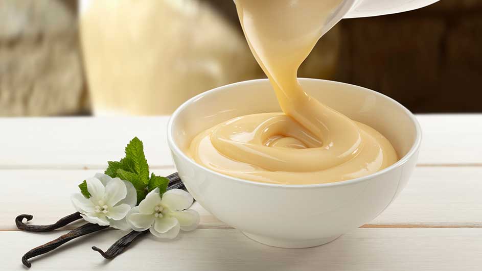 Un bol blanc rempli de sauce jaune est présenté avec des gousses de vanille, des fleurs blanches et de la menthe.