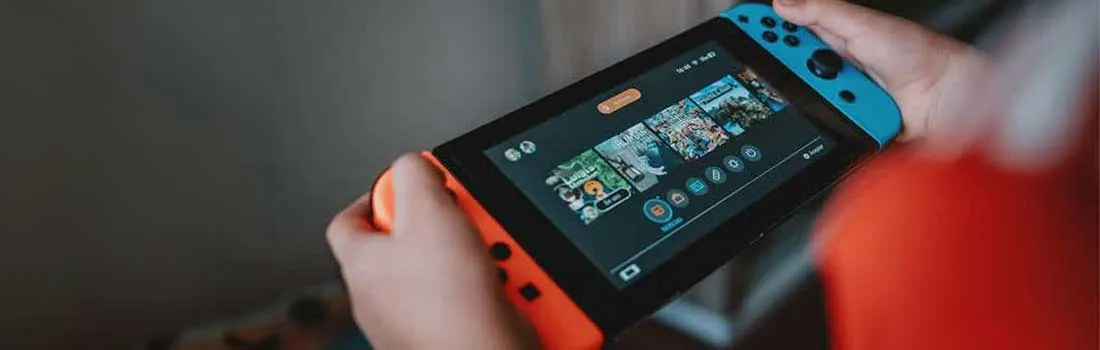 Hands hold a Nintendo Switch with a game menu on its black screen. The Joy-Cons are blue and orange. The background is gray.