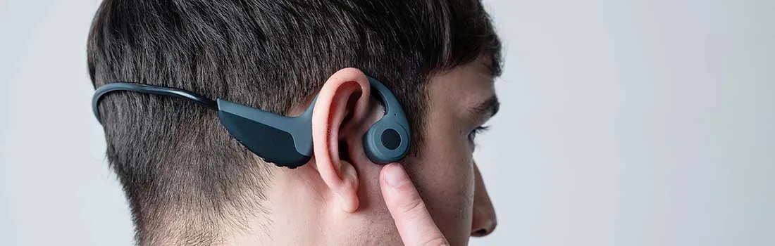 Un homme porte des écouteurs gris et noirs autour de l'oreille. Un doigt touche l'écouteur sur fond blanc.