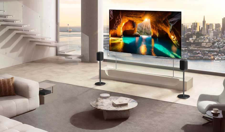 In een moderne woonkamer staat een grote tv met een grotscène; naast het scherm staan speakers en buiten ligt een stadszicht.