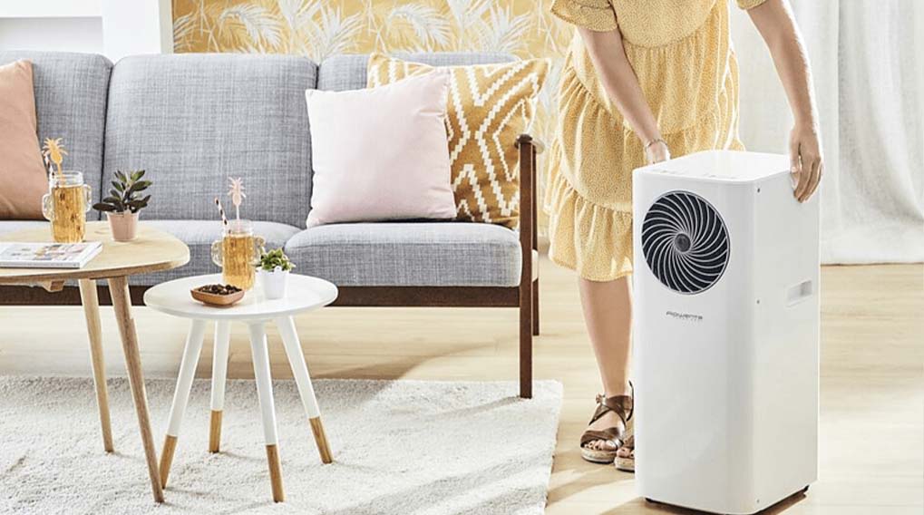 Een witte mobiele airconditioner staat in een kamer met een grijze sofa, houten tafels en decoratieve kussens.