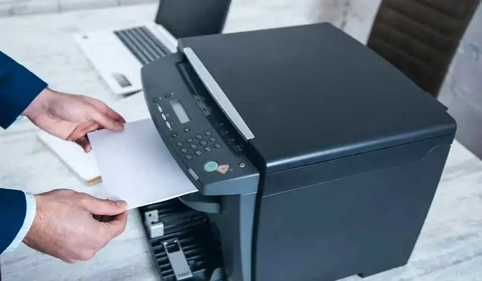 Een donkergrijze printer op een bureau, met een hand die papier in de invoer plaatst. Op de achtergrond staat een laptop.