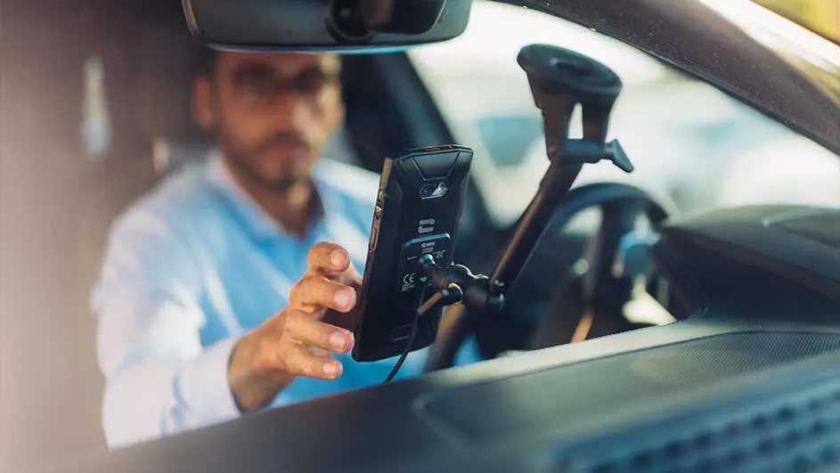 Dans une voiture, un smartphone noir est fixé sur un support. Une personne attrapant et le téléphone et un volant est visible en arrière-plan.