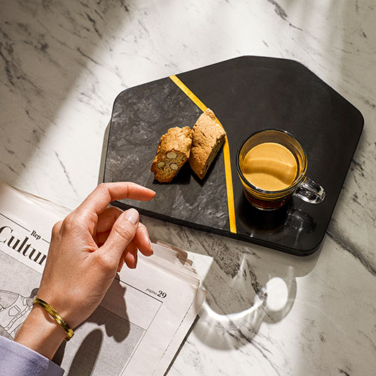 Un plateau noir avec café, biscuits et une bande jaune sur un fond marbré, à côté d'un journal.