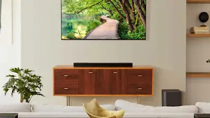 Een woonkamer toont een tv met een bospad, een houten kast met een soundbar, en een witte bank met een gouden schaal.