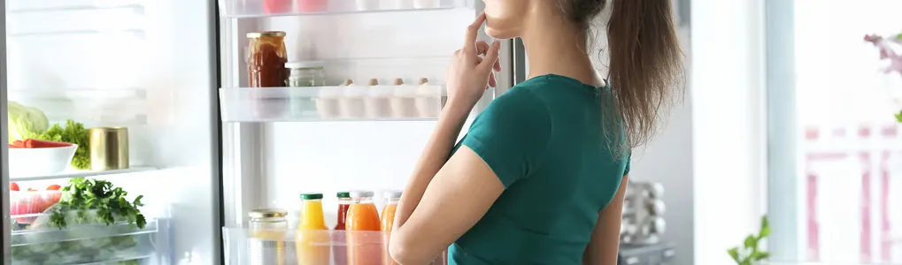Un réfrigérateur blanc ouvert montre des légumes, des fruits et des jus. Des bocaux en verre sont visibles sur l'étagère du haut.