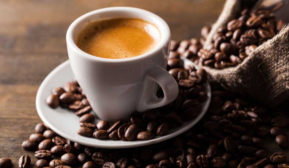 A white cup of espresso sits on a saucer surrounded by coffee beans. In the background, a jute sack filled with more beans rests on a wooden table.