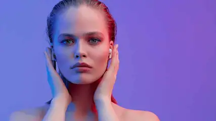 Une personne avec un écouteur blanc tient ses mains près de ses oreilles sur un fond violet.