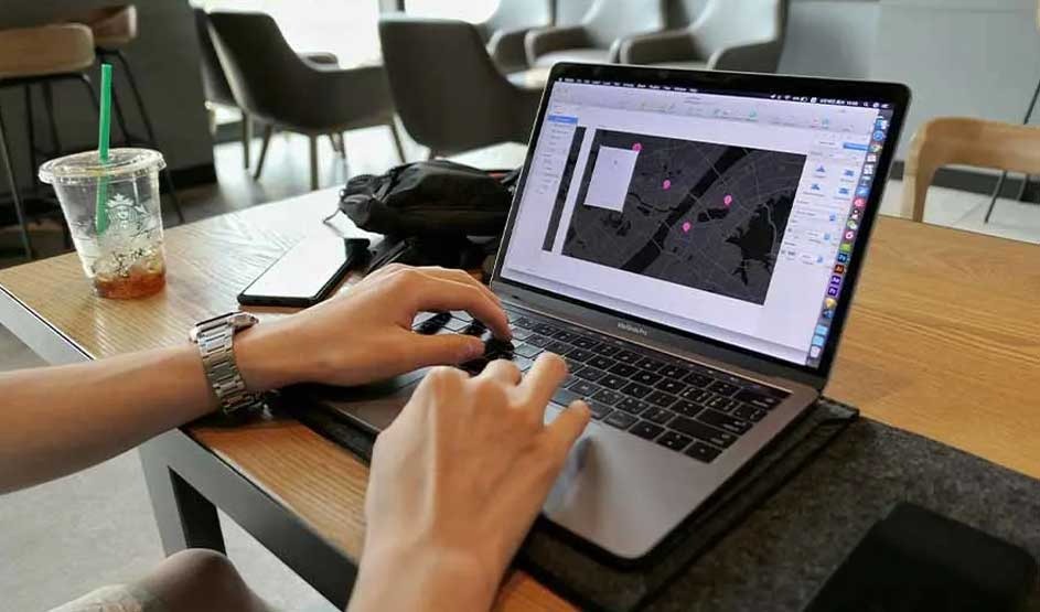 Hands work on an Apple MacBook in a café, map pins on screen, with a phone, bag, and a Starbucks iced drink on the wooden table.
